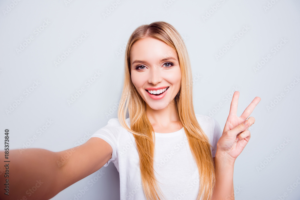 Cute charming lovely positive attractive young lady with blonde hair dressed in white t-shirt is taking a self portrait and demonstrating v-sign, isolated on grey background Cute charming lovely positive attractive young lady with blonde hair dressed in white t-shirt is taking a self portrait and demonstrating v-sign, isolated on grey background