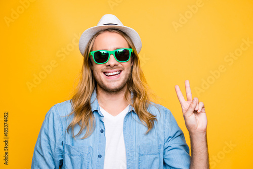 Portrait of happy, attractive, bearded guy in summer glasses with beaming smile showing v-sign in camera standing over yellow background