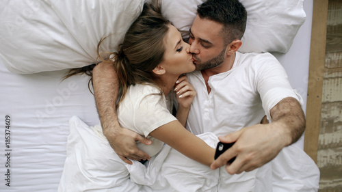 Young beautiful and loving couple take selfie picture on smartphone camera and kiss while lying in bed at the morning