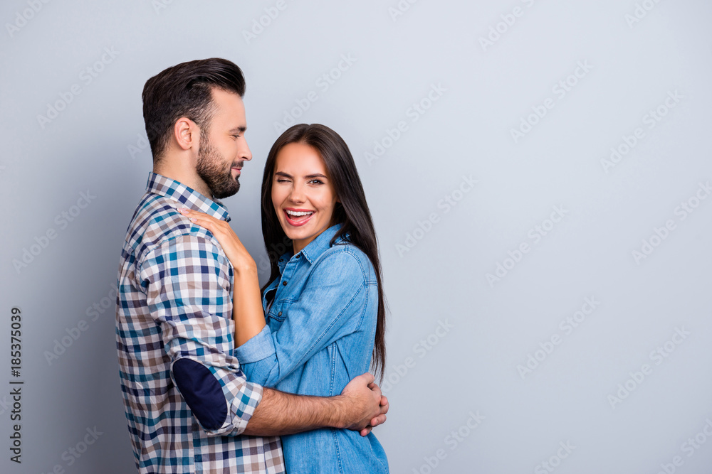 Portrait of caucasian, attractive couple in shirts - man with bristle embrace his charming, funny, pretty woman, who blink to the camera over grey background Portrait of caucasian, attractive couple in shirts - man with bristle embrace his charming, funny, pretty woman, who blink to the camera over grey background