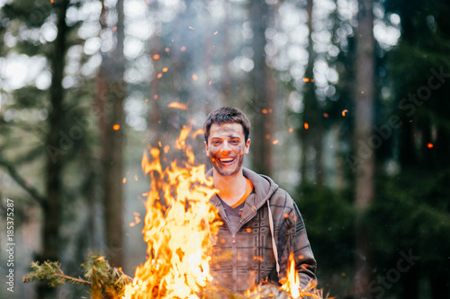 Happy funny crazy fearless man holding burning firewood in hands. Excited bizarre unusual boy laughing with stained dirty face in forest at nature. Fire in travel camp. Young male playing with flame. photo