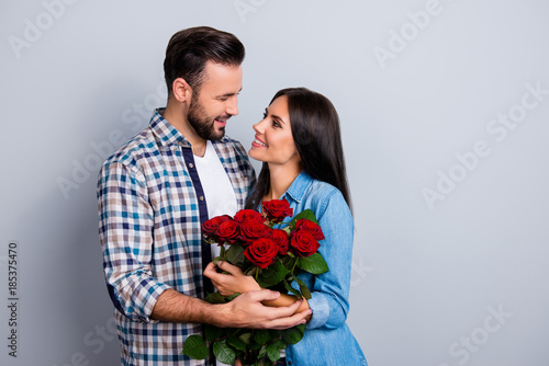 Beautiful, happy, positive couple embracing, looking to each other, holding bouquet of red roses over grey background, 14 february, young, cute family going to be parents