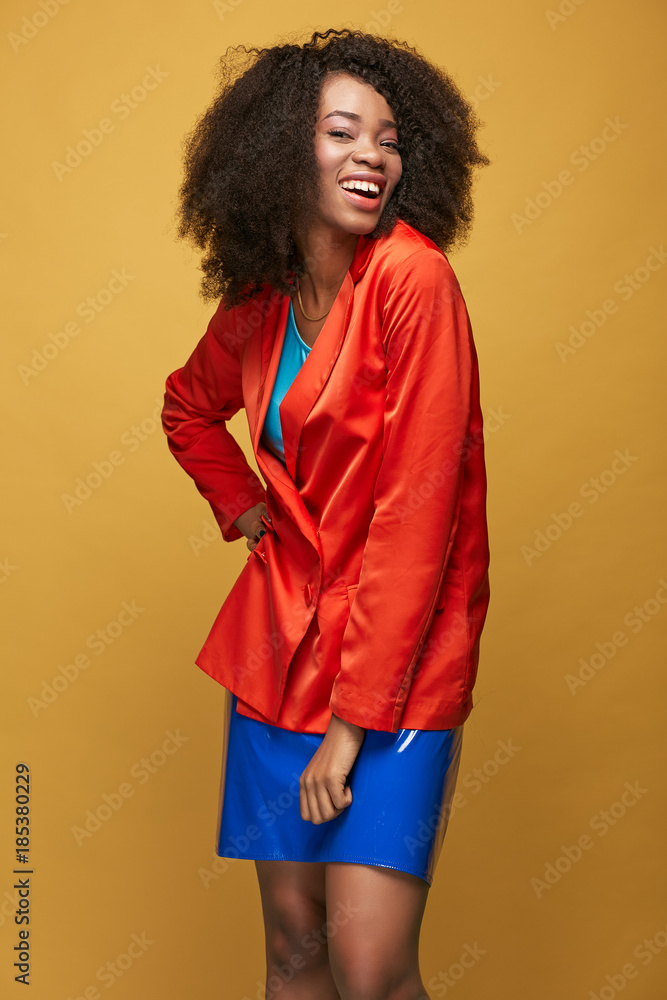 Beauty portrait of attractive young african girl with afro hairstyle. Amazing girl wearing orange red jacket, blue latex skirt posing on yellow background. Studio shot. Beauty portrait of attractive young african girl with afro hairstyle. Amazing girl wearing orange red jacket, blue latex skirt posing on yellow background. Studio shot.