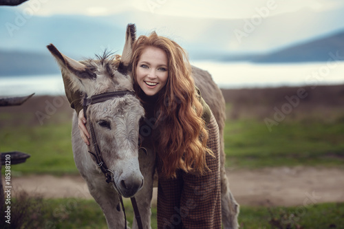 A girl with curly red hair in fashionable clothes in the style of Provence hugs a cute donkey photo