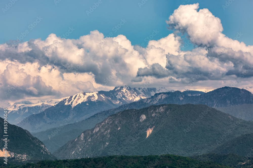 The view of the Caucasus mountains covered with green forest and snow under large cumulus clouds from the lookout tower on mount Akhun. Khosta district, Adler, Sochi, Russia.