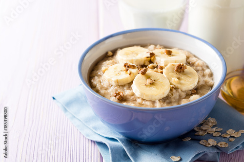 Oatmeal porridge with banana, walnuts and honey in bowl on purple wooden background. Healthy breakfast.