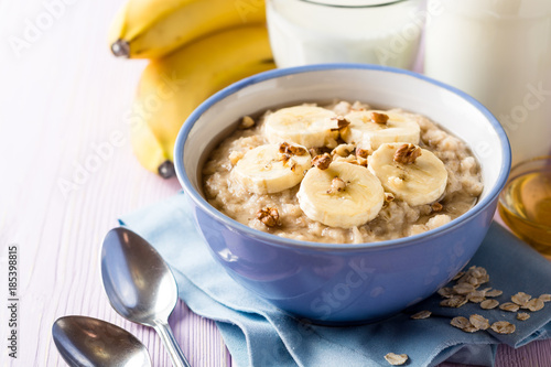 Oatmeal porridge with banana, walnuts and honey in bowl on purple wooden background. Healthy breakfast.