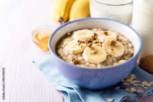 Oatmeal porridge with banana, walnuts and honey in bowl on purple wooden background. Healthy breakfast.