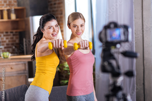 Sports equipment. Positive cheerful well built women smiling and looking into the camera while working out with dumbbells