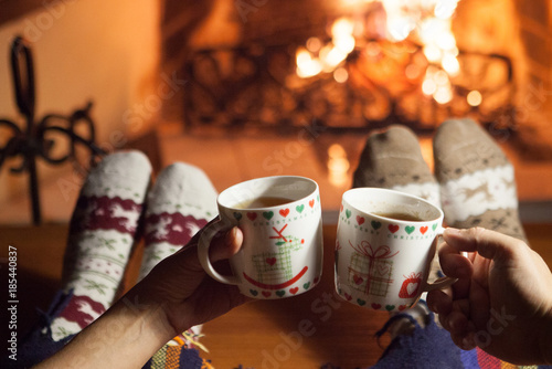 Man and woman in warm knitted socks with cups of hot drink in front of the fireplace