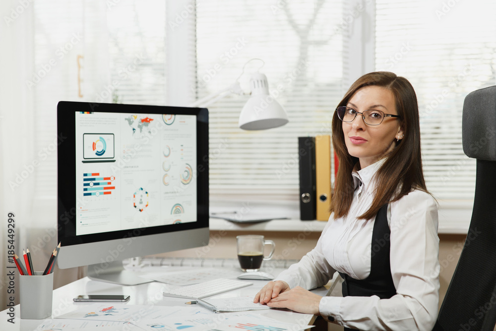 Beautiful smiling brown-hair business woman in suit and glasses sitting at the desk with cup of coffee, working at computer with documents in light office, looking at the camera Beautiful smiling brown-hair business woman in suit and glasses sitting at the desk with cup of coffee, working at computer with documents in light office, looking at the camera