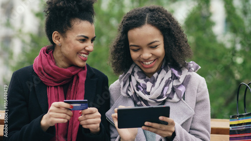 Two attractive mixed race women with paper bags sitting on bench and using tablet computer and credit card for online shopping