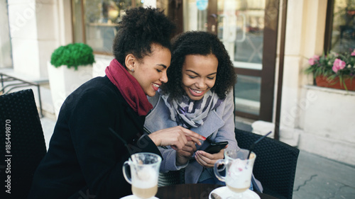 Two attractive mixed race female friends sharing together using smartphone in street cafe outdoors photo