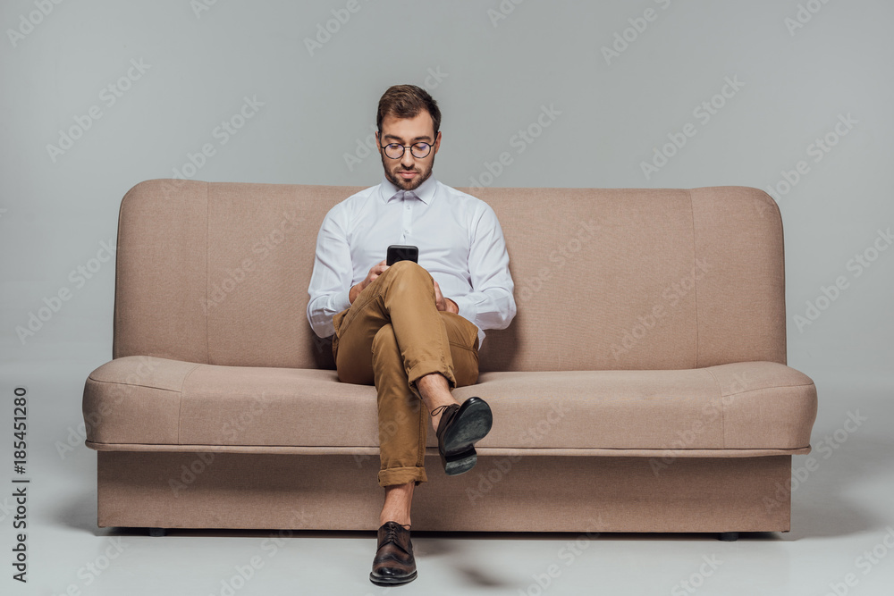 stylish man in eyeglasses sitting on couch and using smarttphone isolated on grey - obrazy, fototapety, plakaty 