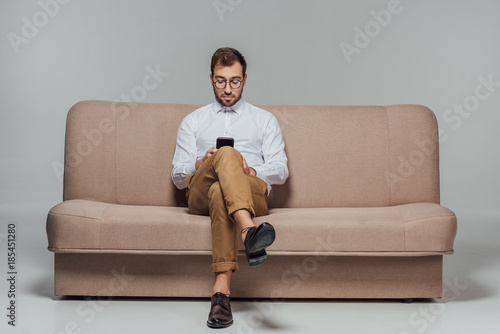 stylish man in eyeglasses sitting on couch and using smarttphone isolated on grey