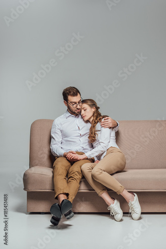 handsome young man hugging beautiful girlfriend sleeping on his shoulder while sitting on sofa isolated on grey