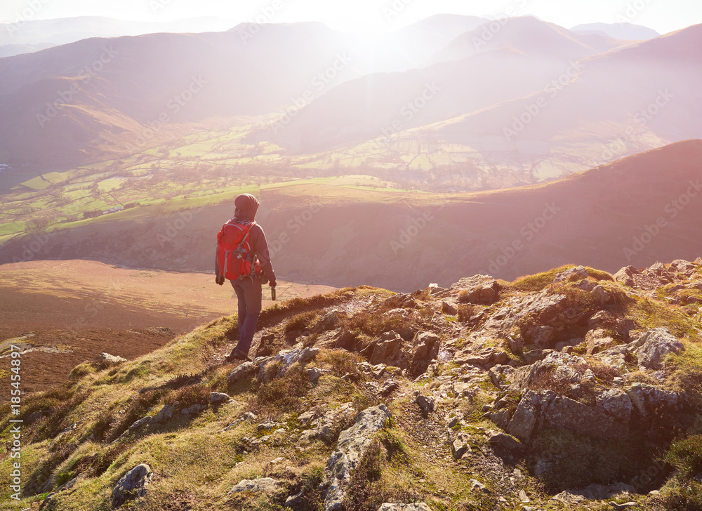 A hiker and their dog walking down the mountain ridge of Scar Crags and Causey Pike with the early morning sun in the English Lake District, UK.