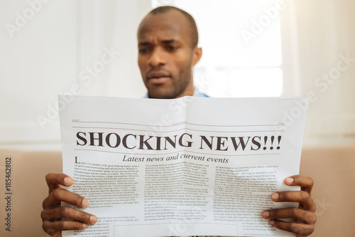 Being astonished. Serious astonished afro-american man holding a newspaper and reading shocking news while sitting on the sofa