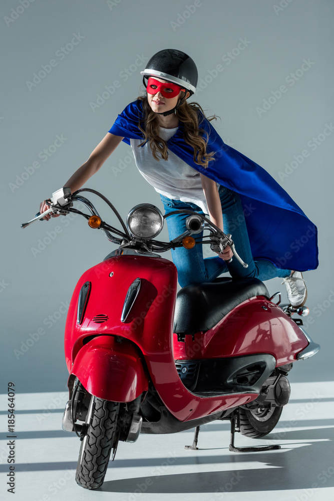 woman in superhero mask and cape and protective helmet riding red scooter woman in superhero mask and cape and protective helmet riding red scooter