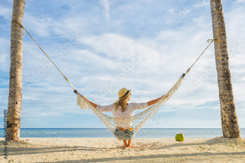 Woman in hat sitting in hammock on the beach. Travel and vacation concept.