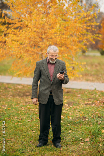 A handsome elderly man wearing glasses is using a phone. Walk in the park in autumn