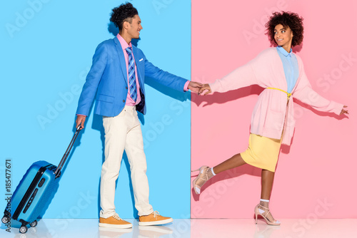 side view of african american couple with suitcase walking on pink and blue wall background