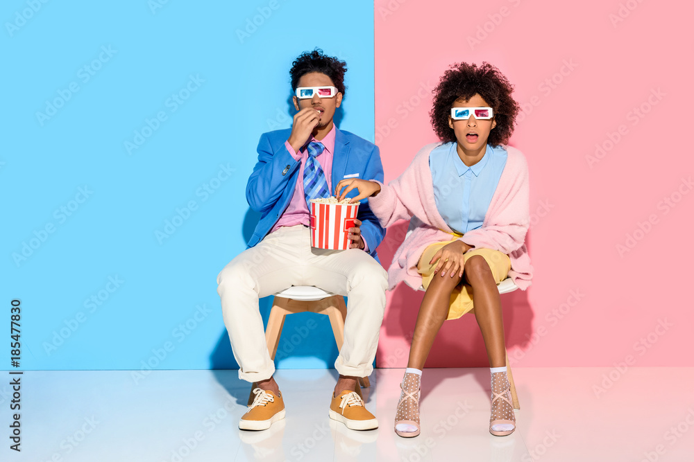 Obraz premium young african american couple sitting on chairs and eating popcorn against pink and blue wall