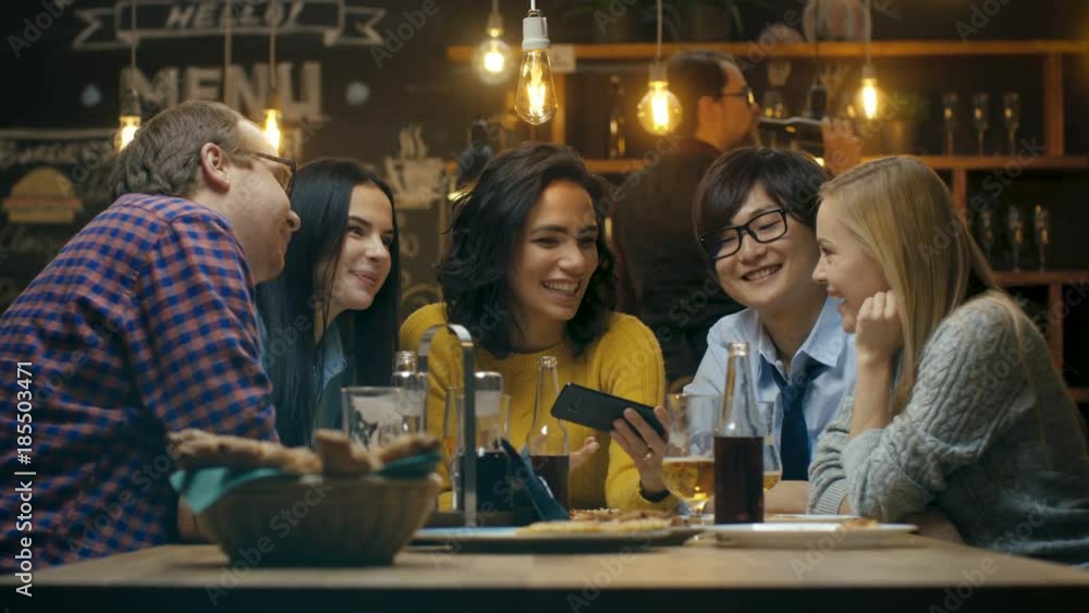 Beautiful Hispanic Woman Shows Interesting Stuff on Her Smartphone to Her Friends while They Have Good Time in Bar. They Laugh, Joke, Drink in Stylish Hipster Bar Establishment. Beautiful Hispanic Woman Shows Interesting Stuff on Her Smartphone to Her Friends while They Have Good Time in Bar. They Laugh, Joke, Drink in Stylish Hipster Bar Establishment.