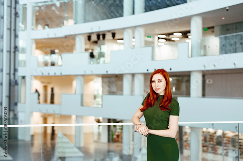 Business portrait of the beautiful red-haired girl in a green dress against the background of office.