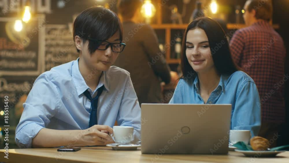 Handsome Asian Man and Beautiful Caucasian Young Woman Sitting in the Cafe Work on a Laptop Computer