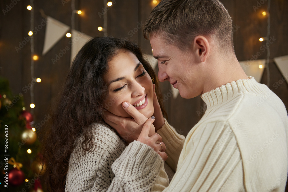 Fototapeta premium Young couple hugging in dark wooden interior with lights. Romantic evening and love concept. New year holiday. Christmas lights and decoration. Dressed in white.