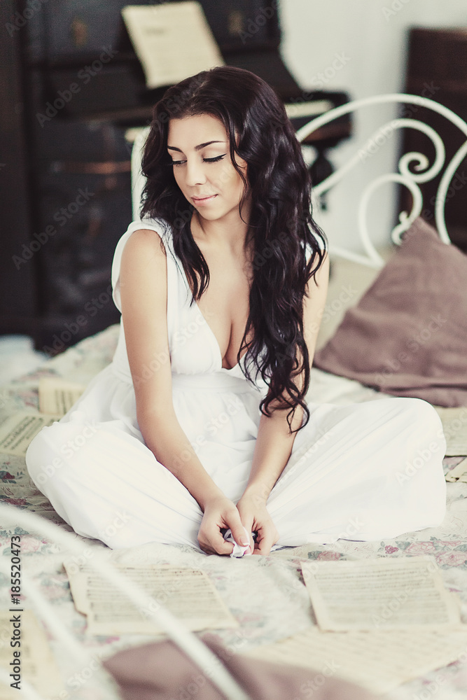 Fototapeta premium Portrait of beautiful woman, sitting on white bed with blurred piano on background