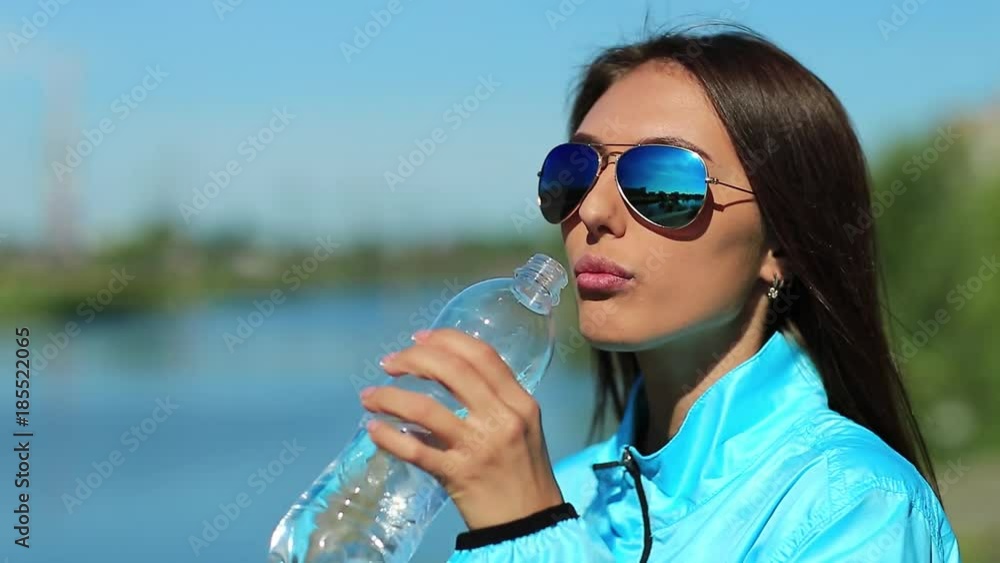 Sportswoman drinks water. Attractive girl in sunglasses drinks clean mineral water from bottle. Beautiful woman in blue sports jacket drinks water and looks around Sportswoman drinks water. Attractive girl in sunglasses drinks clean mineral water from bottle. Beautiful woman in blue sports jacket drinks water and looks around