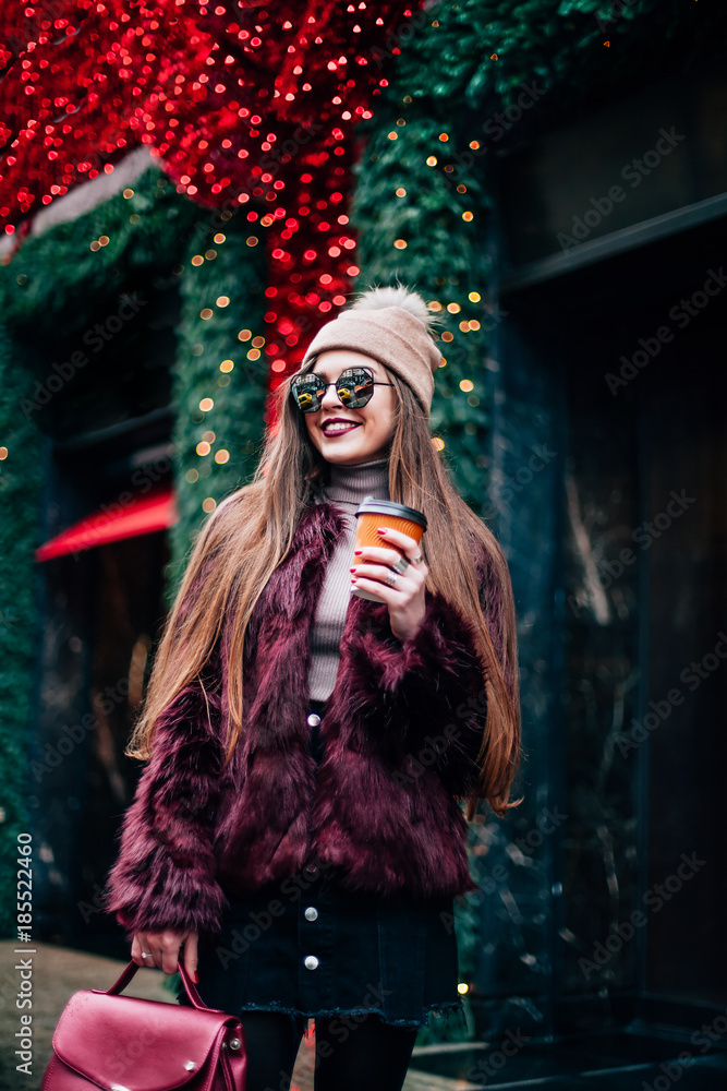 Fototapeta premium Outdoor portrait of young beautiful happy smiling girl posing on street. Model wearing stylish warm clothes.Keeps coffee. concept of street fashion