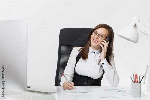 Pretty smiling business woman in suit sitting at the desk, working at contemporary computer with document in light office, talking on mobile phone, conducting pleasant conversation on white background
