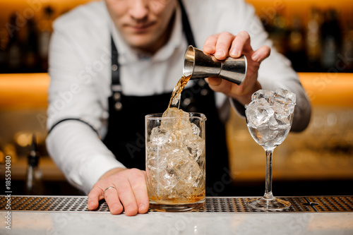 Barman dressed in a white shirt pouring alcoholic drink into a glass with ice cubes