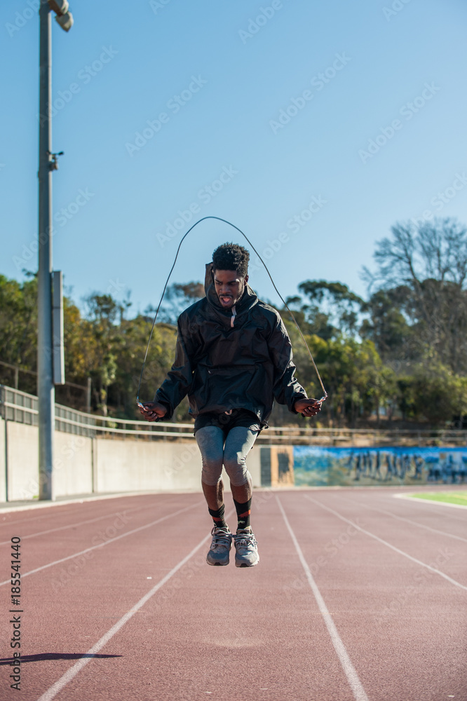 Lean Jamaican athlete jumping rope with bent knees on track at stadium.  