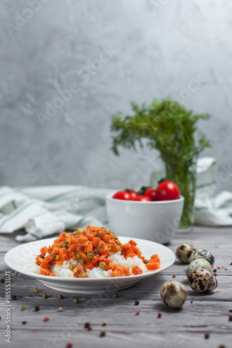 delicious rice with stewed carrots, peas and onions on a white plate, quail eggs, bowl with cherry tomatoes, towel, pepper and dill on a light wooden table