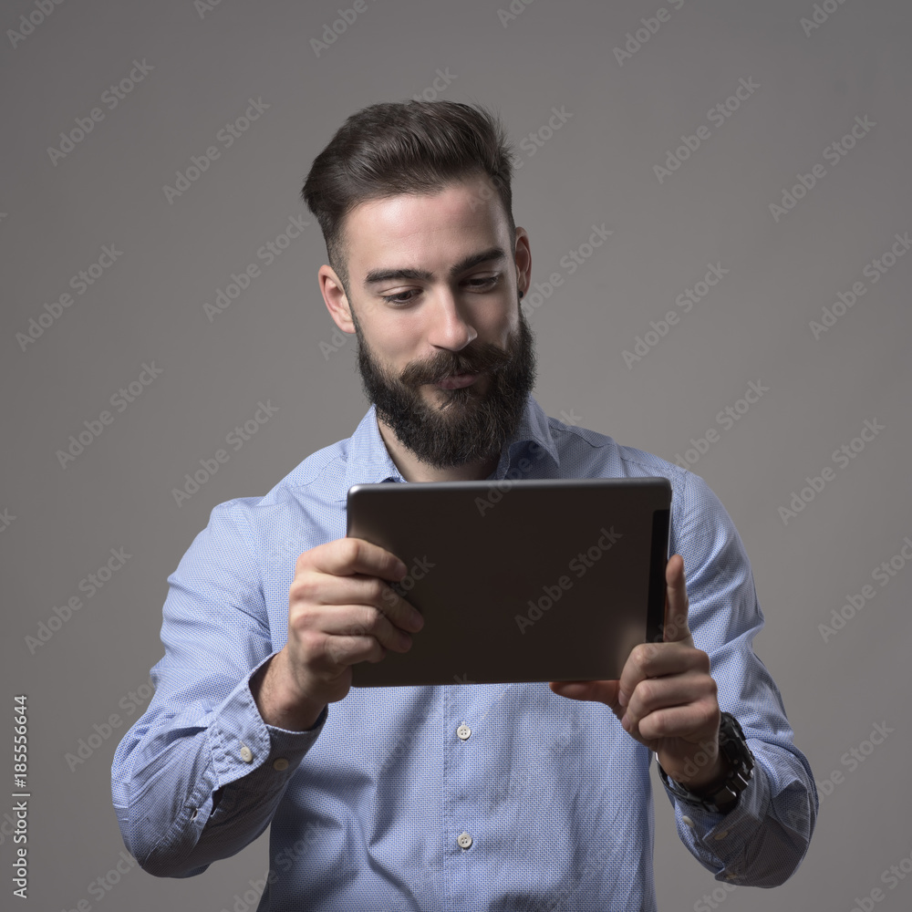 Young happy bearded man holding and reading e-book from touch pad tablet computer over gray studio background. Young happy bearded man holding and reading e-book from touch pad tablet computer over gray studio background.