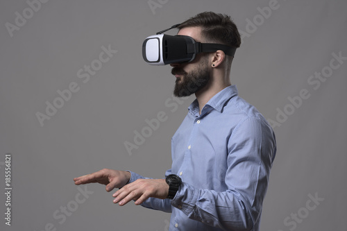 Young bearded businessman with vr glasses typing on augmented reality keyboard simulator over gray studio background. 