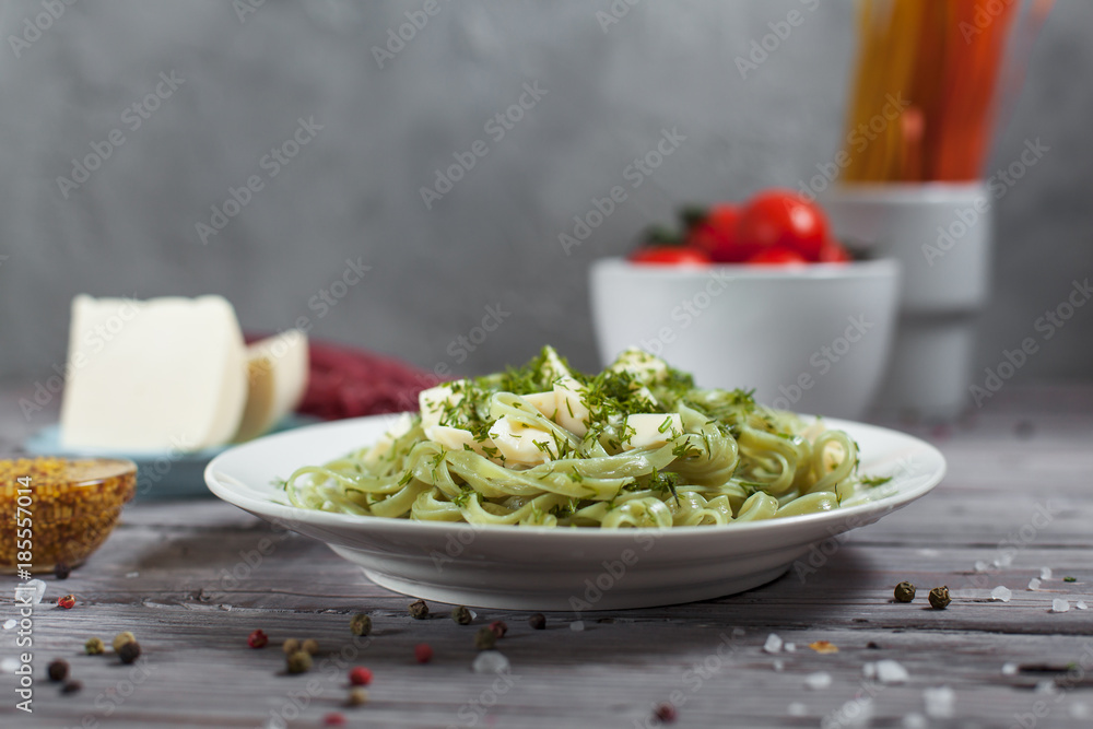 pasta green with spinach and mozzarella cheese and greens in a white plate, French mustard, colored raw spaghetti, saucer with cheese, dill and bowl with cherry tomatoes on a light wooden table