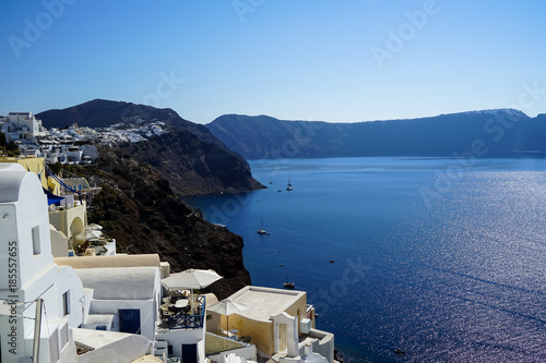 Panoramic view of blue Aegean sea, sailing ships and ocean water reflection from Oia village with white buildings cityscape along island mountain and blue sky background