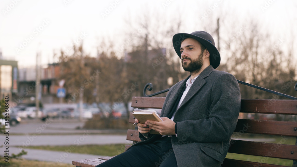 Creative young writer man in hat and coat write notes for his future book with pen in notebook sitting on city street bench at park Creative young writer man in hat and coat write notes for his future book with pen in notebook sitting on city street bench at park