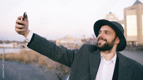 Young happy tourist man in ahat and coat smiling while taking selfie picture with mobile phone on city riverside photo