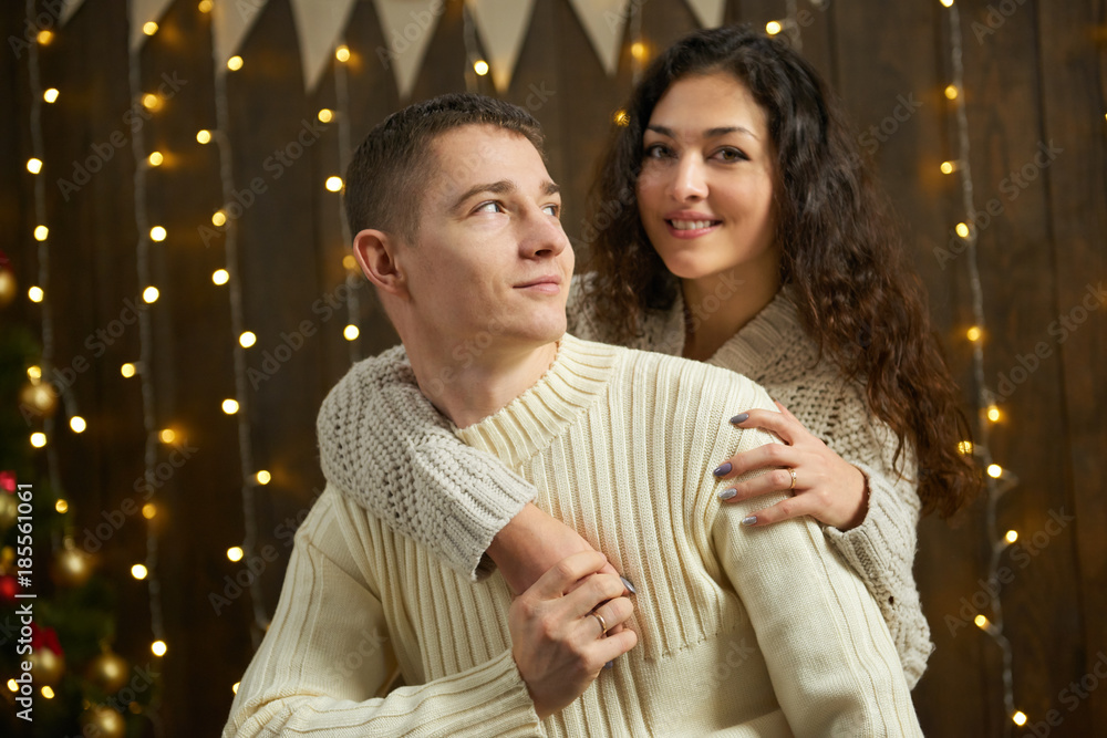 Fototapeta premium couple in christmas lights and decoration, dressed in white, young girl and man, fir tree on dark wooden background, winter holiday concept