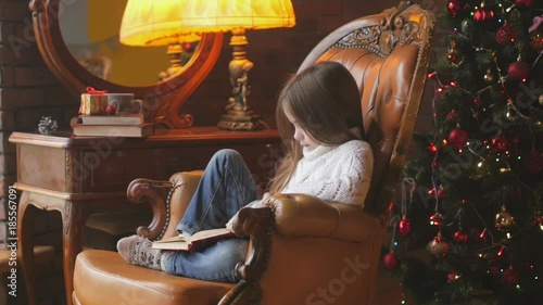 beautiful little girl is reading a book while sitting in a chair near a festive Christmas tree