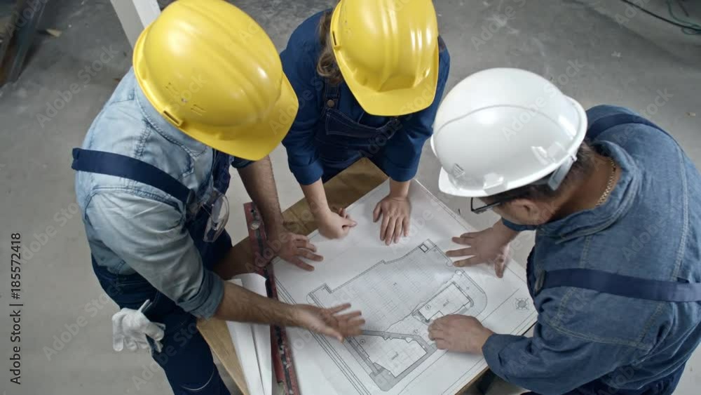 Directly above shot of male and female construction workers in hard hats standing and discussing floor plan draft