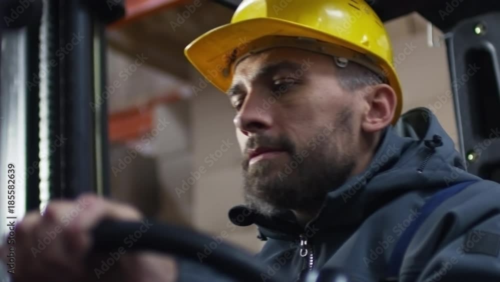 Low angle shot of bearded male worker in hard hat operating forklift in warehouse Low angle shot of bearded male worker in hard hat operating forklift in warehouse