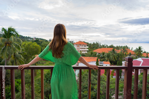A young adult woman in green dress is standing on a balcony of luxury hotel in tropical country and looking forward on sea and feel freedom and harmony