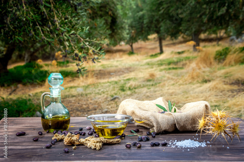 Bowl and bottle with extra virgin olive oil, olives, a fresh branch of olive tree and cretan rusk dakos on wooden table, in an olive tree field at Crete, Greece.